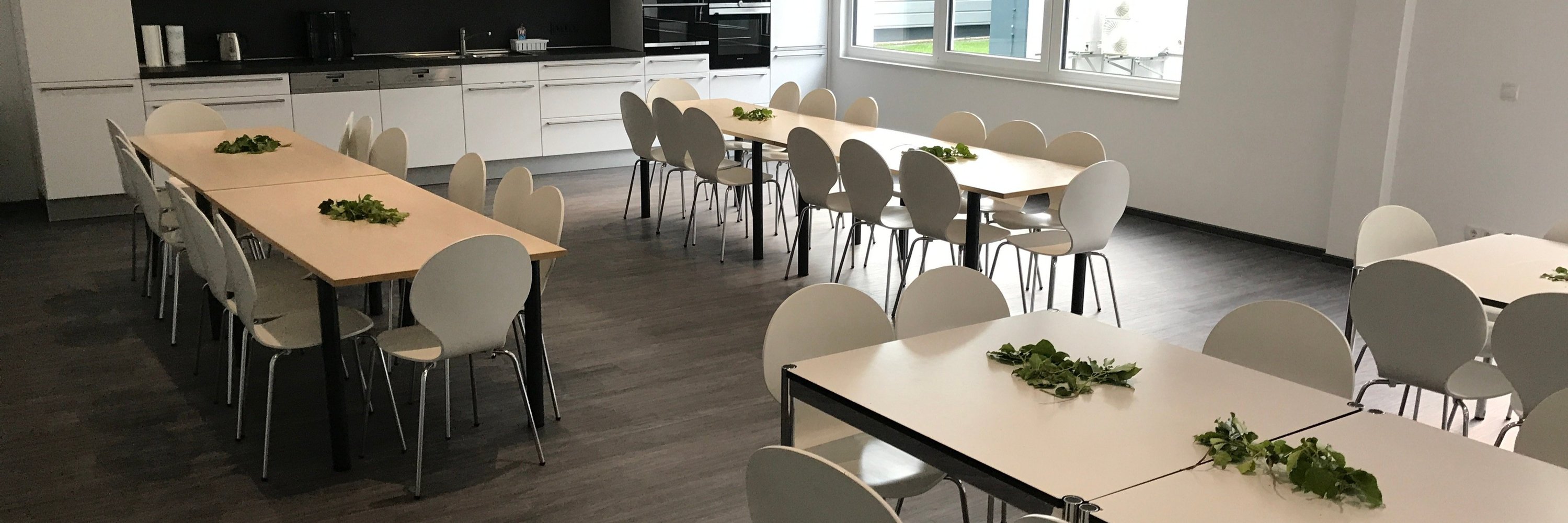 Moderner großer Aufenthaltsraum mit weißer Einbauküche und dunklem Bodenbelag in Holzoptik.